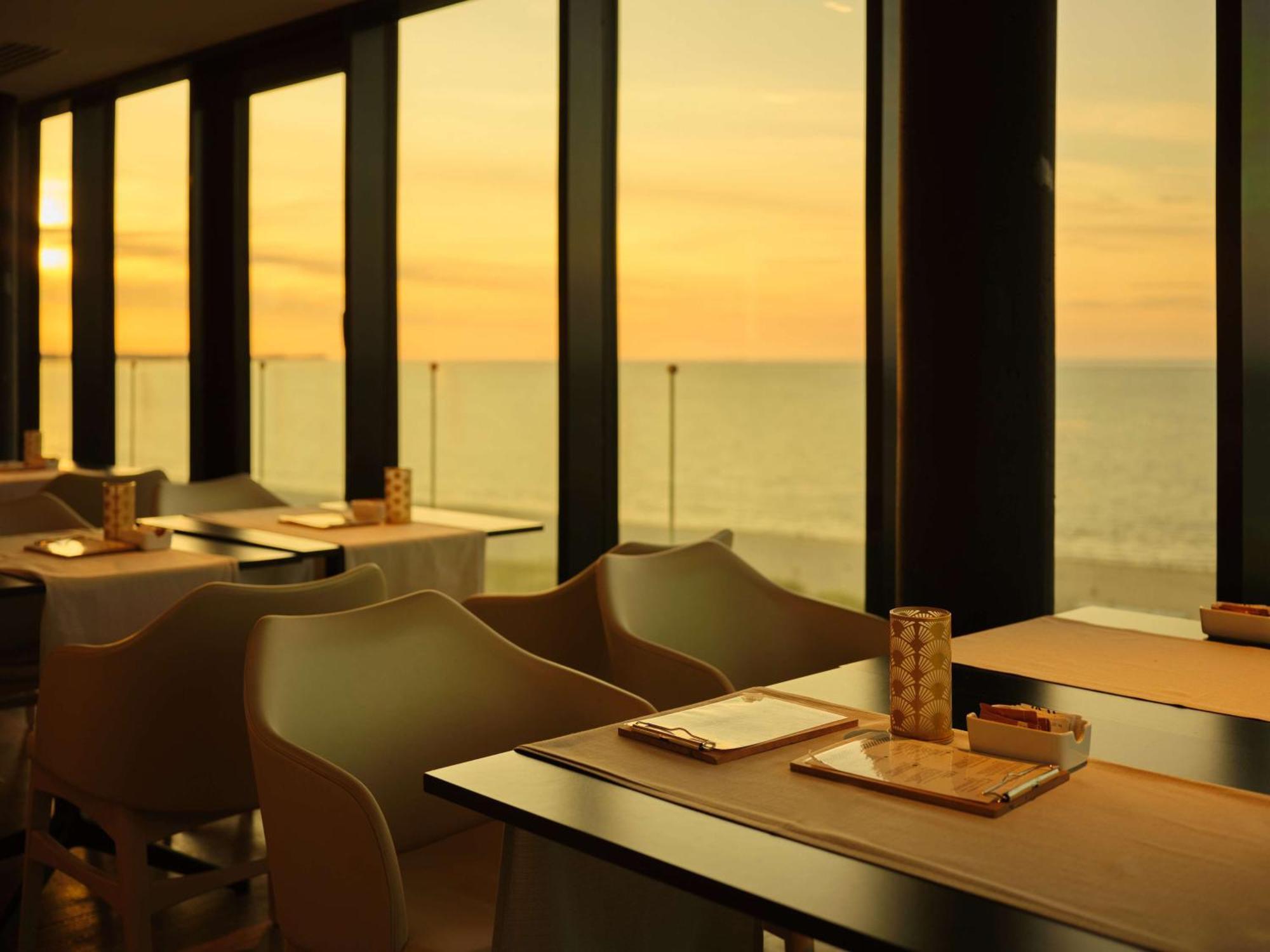 Radisson Blu Resort Swinoujscie Bagian luar foto The photo shows a restaurant interior with a beautiful view of the ocean during sunset. The large windows allow natural light to flood the space, highlighting the warm colors of the sunset in the sky. There are several tables, each set with placemats