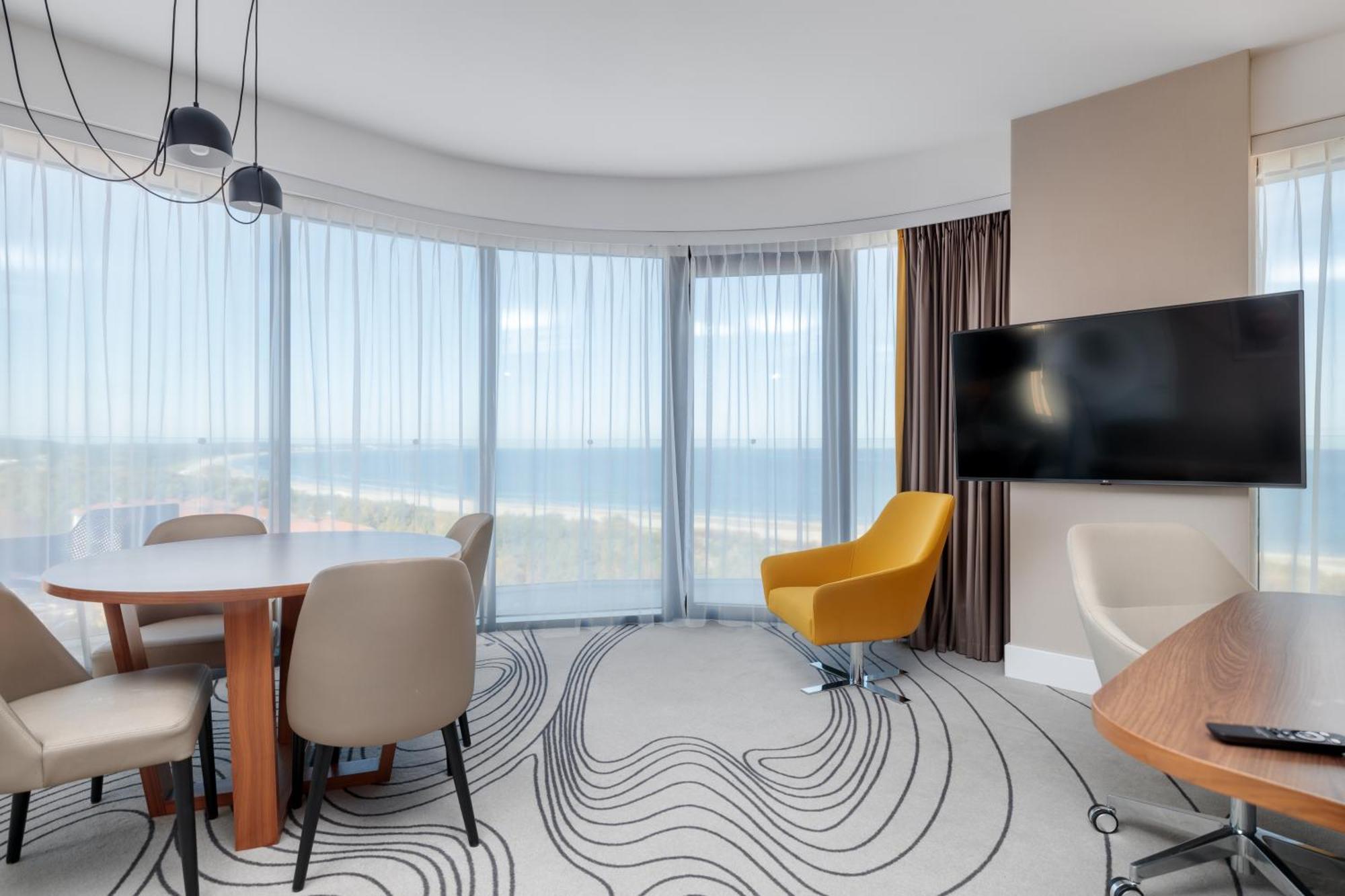 Radisson Blu Resort Swinoujscie Bagian luar foto The image shows a modern hotel room or suite with a bright, airy atmosphere. There are large windows that allow natural light to fill the space, providing a view of the ocean. The decor includes a round dining table with several chairs, and a stylish