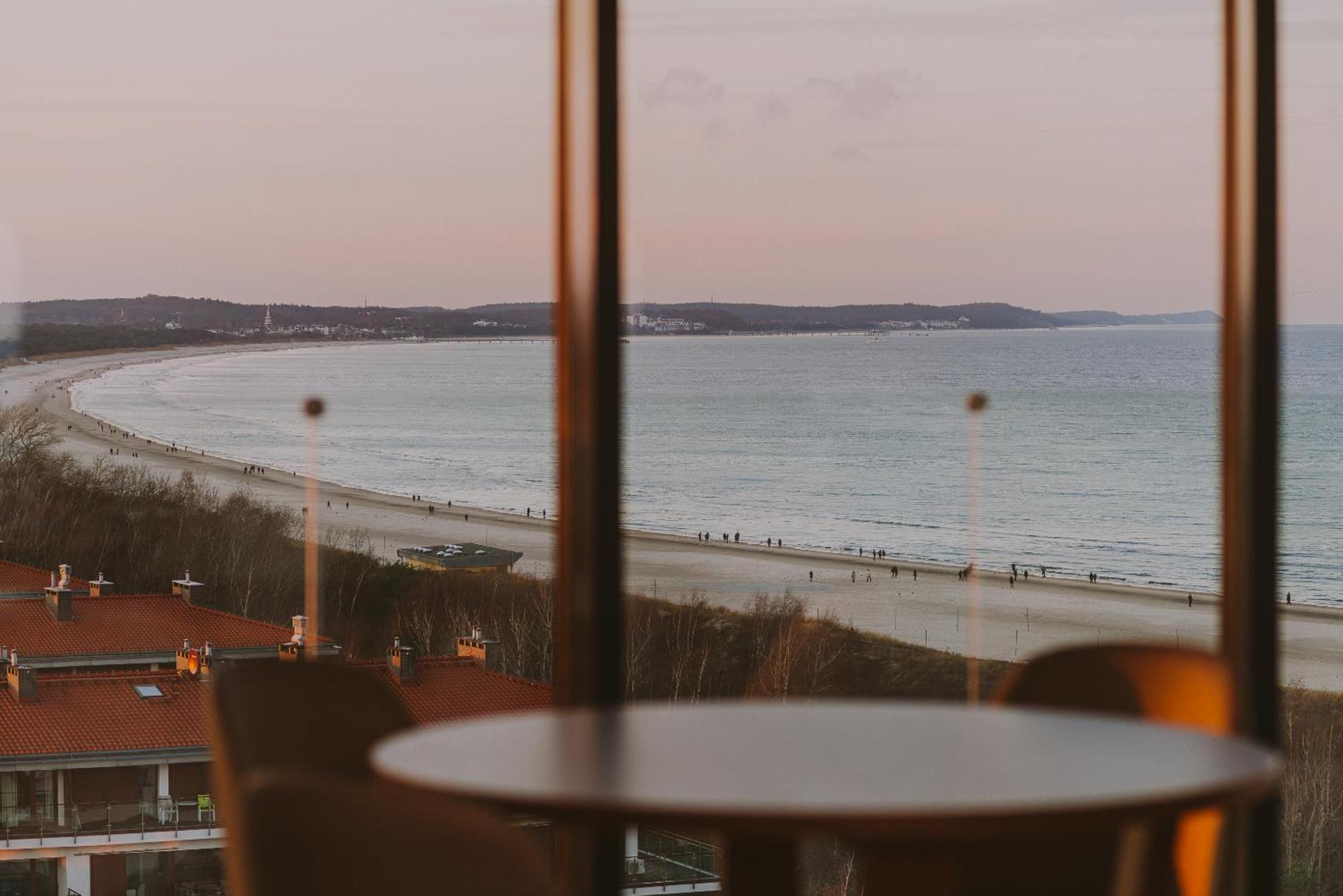 Radisson Blu Resort Swinoujscie Bagian luar foto The photo shows a picturesque view of a beach scene from indoors. In the foreground, there is a round table with some chairs around it, suggesting a cozy interior space. Through a large window, you can see the beach with gentle waves hitting the shor