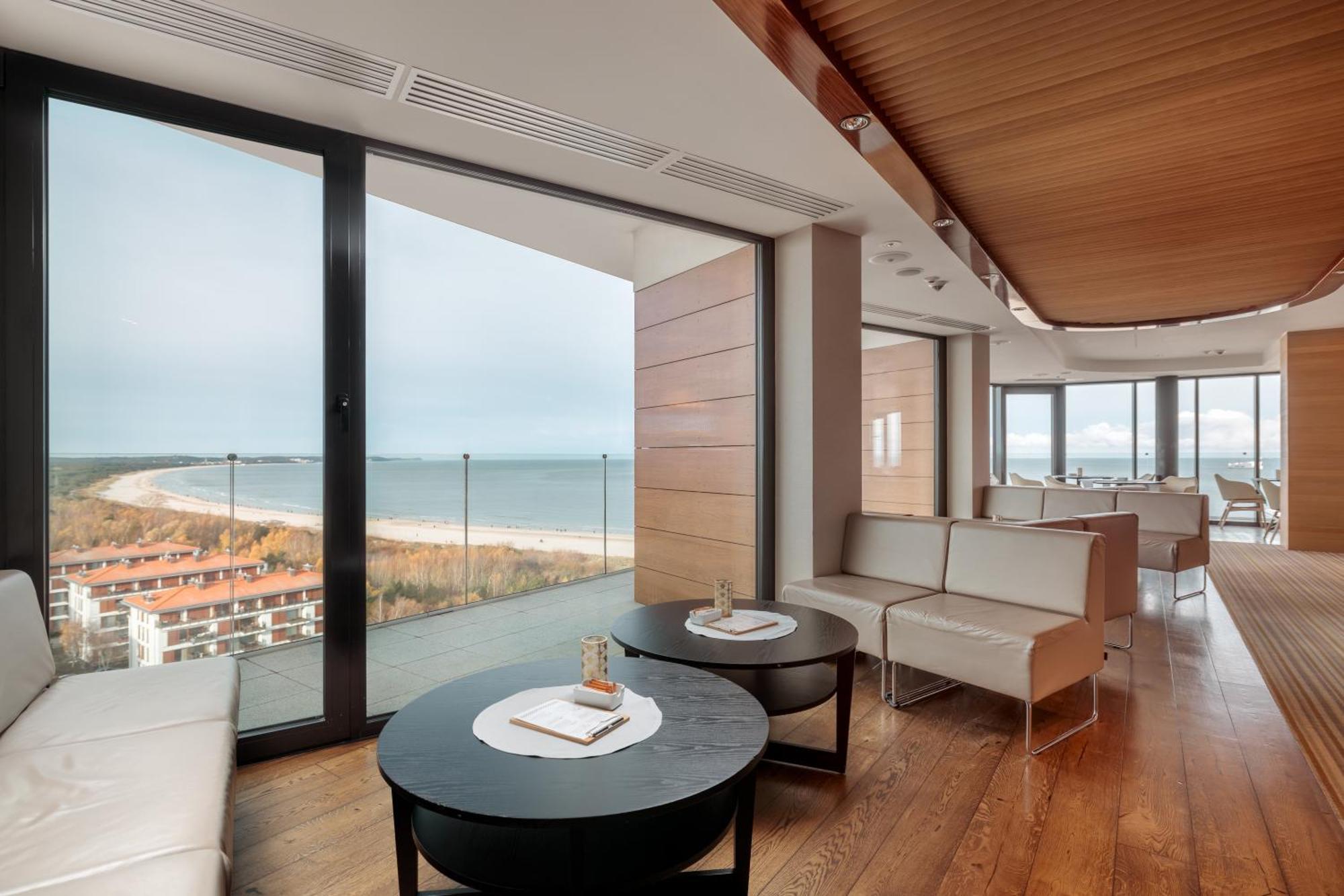Radisson Blu Resort Swinoujscie Bagian luar foto The photo shows a modern, elegantly designed interior space featuring large windows that offer a panoramic view of the ocean and coastline. The flooring is wooden, providing a warm atmosphere. There are two round black tables in the foreground with m