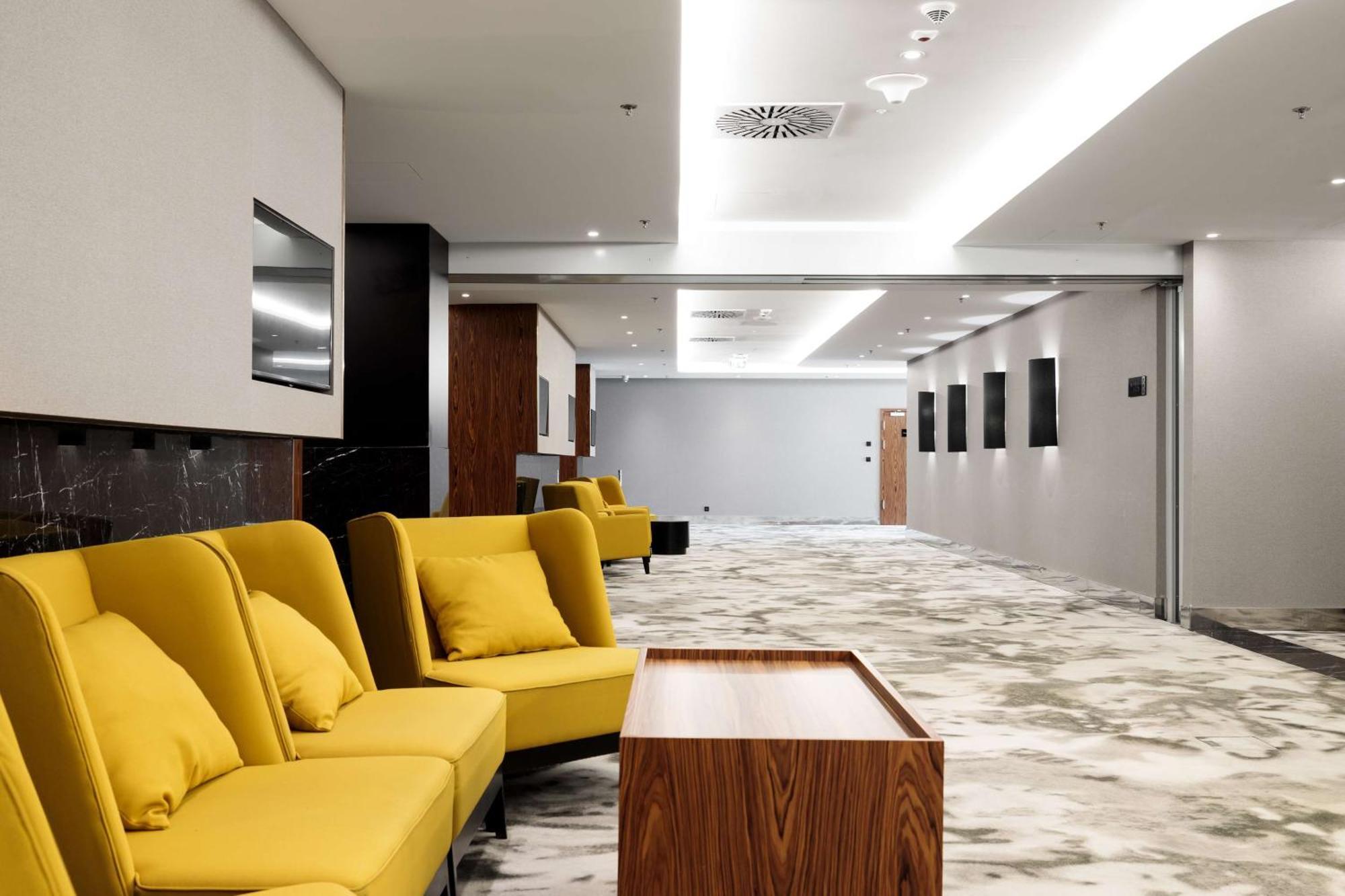Radisson Blu Resort Swinoujscie Bagian luar foto The photo shows a modern interior space, likely a lobby or waiting area. There are several yellow sofas with cushions arranged in a casual seating area. In front of the sofas, there is a wooden coffee table. The flooring features a gray and white mar