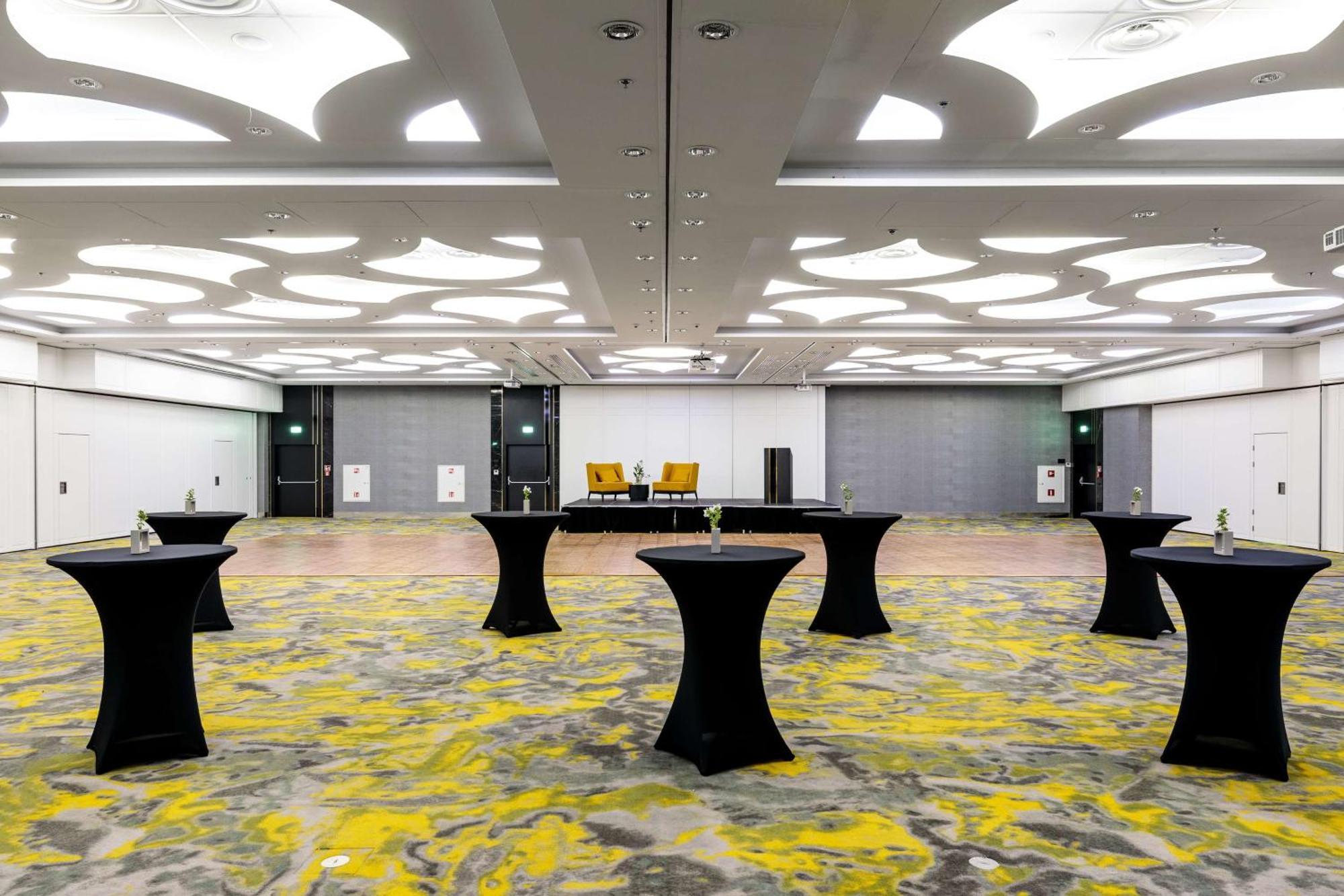 Radisson Blu Resort Swinoujscie Bagian luar foto The photo shows a spacious interior room set up for an event. The flooring features a patterned design in gray with yellow accents. There are several black tall tables scattered throughout the area, and in the background, there are two large yellow a