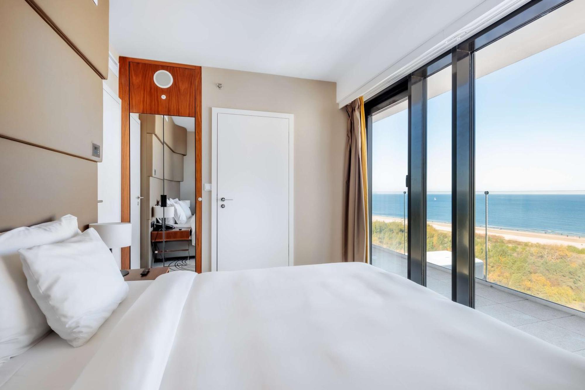 Radisson Blu Resort Swinoujscie Bagian luar foto The photo shows a modern hotel room with a bright and airy atmosphere. There is a large bed with white bedding positioned near a glass wall that offers a view of the ocean. The room features neutral-colored walls and wooden accents, enhancing its con