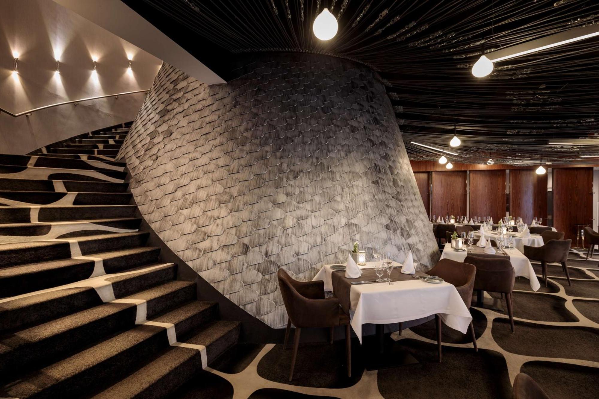 Radisson Blu Resort Swinoujscie Bagian luar foto The photo shows the interior of a stylish restaurant. In the foreground, there are elegantly set tables with white tablecloths and dark chairs. A unique, curving stone wall adds a distinct architectural feature, and a grand staircase with a textured 