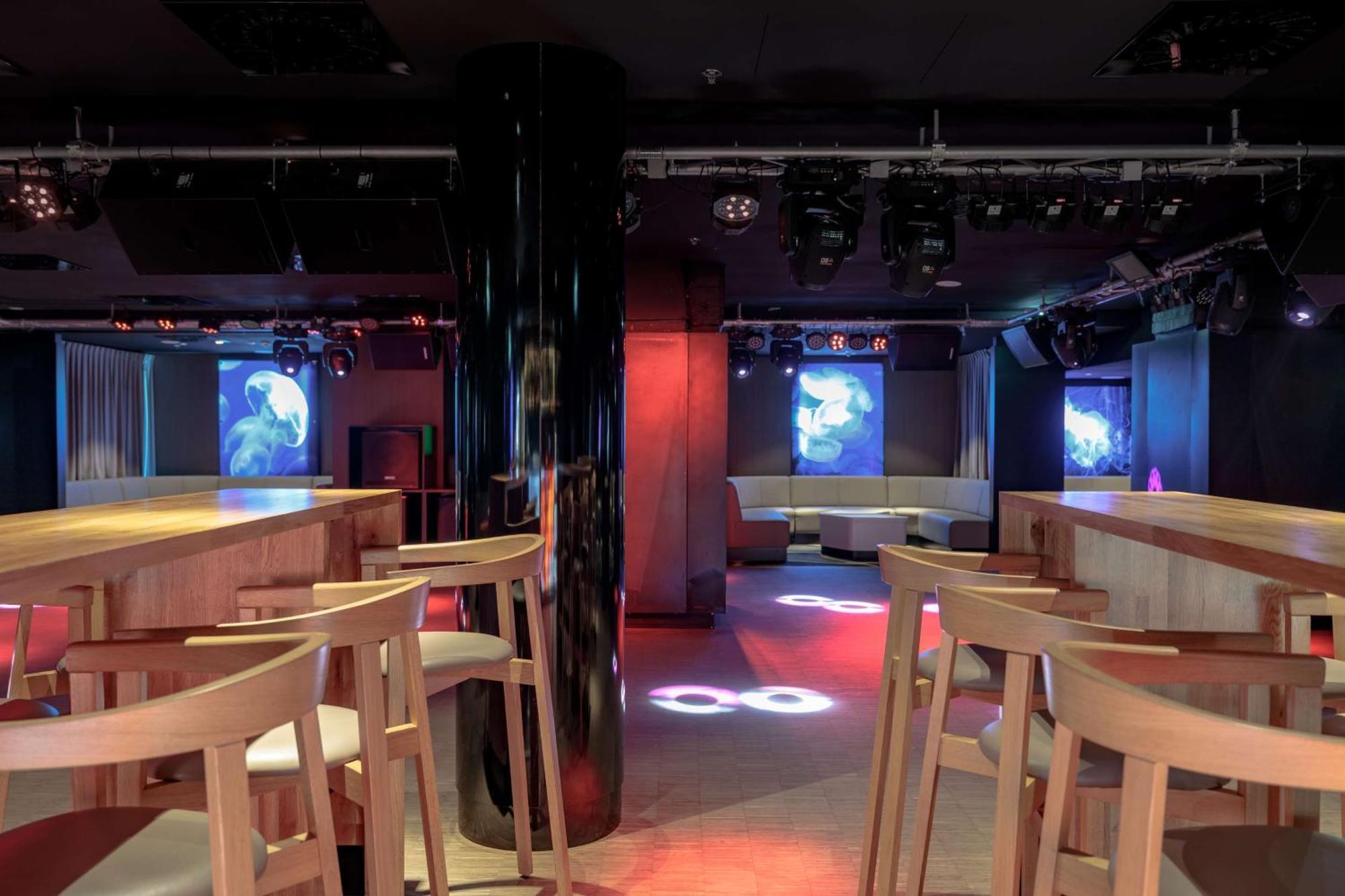 Radisson Blu Resort Swinoujscie Bagian luar foto The photo shows an interior of a modern venue, likely a bar or club. The space features a central black column and light-colored wooden bar stools arranged around a bar area. On the walls, there are large screens displaying vibrant visuals, possibly 
